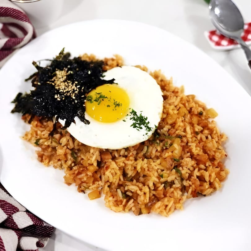 Kimchi fried rice with a sunny-side-up egg, seaweed, and sesame seeds on a white plate.