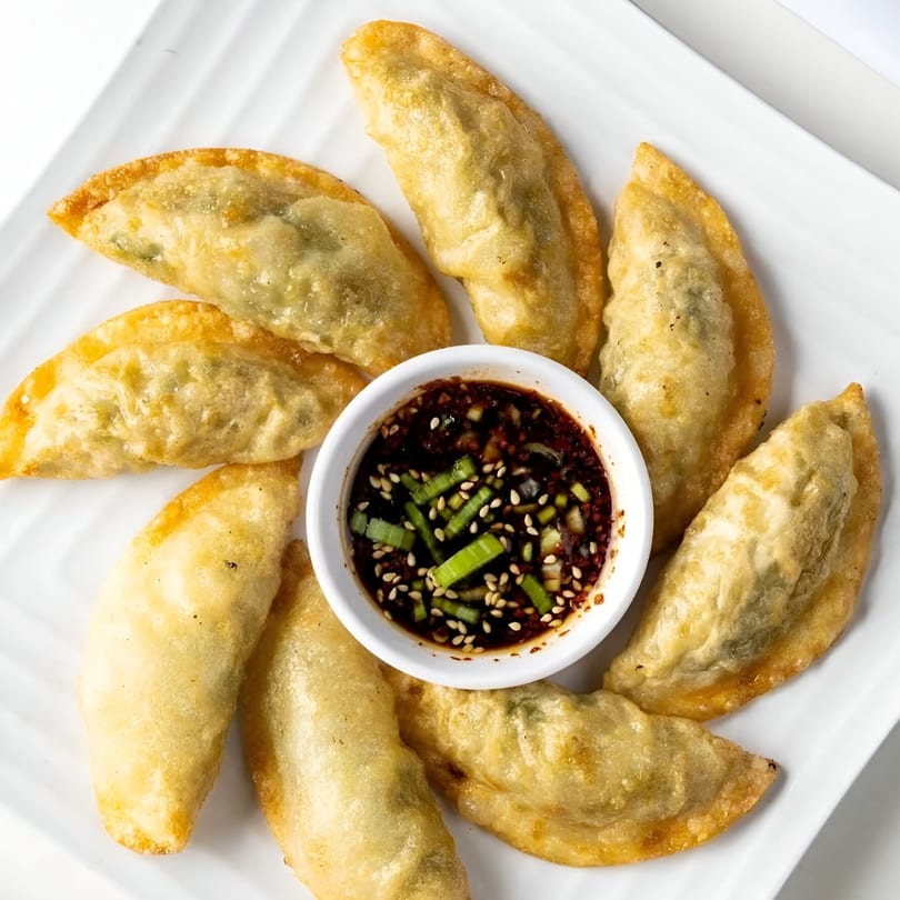 Golden-fried Korean mandu dumplings arranged around a small bowl of soy dipping sauce with scallions and sesame seeds.